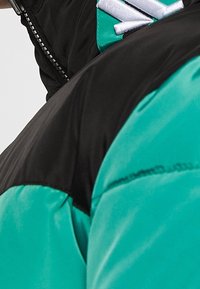 Färgblockerad jacka med en glänsande svart överdel och en livlig turkos nederdel. Inkluderar dragkedja och vita logotypdetaljer. Tyget verkar slätt.