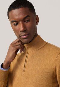 Pull en maille beige avec un col montant et un design à demi-fermeture éclair. Présente des poignets côtelés, et le tissu a une texture lisse. En dessous, une chemise rayée bleue est visible.