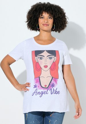Vrouw met krullend haar die een wit T-shirt draagt met een gestileerd vrouwelijk gezicht, een rode hoofddoek en de tekst “Angel Vibe”, gecombineerd met met verf bespatte spijkerbroek.