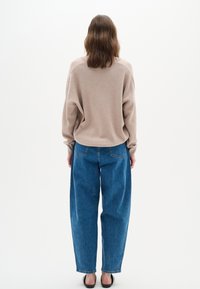 Osoba z brązowymi włosami do ramion, ubrana w beżowy sweter, niebieskie dżinsy i czarne buty wsuwane, stojąca przodem do białego tła.