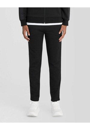Person iført sorte slim-fit bukser, sort jakke, hvid skjorte og hvide sneakers, stående på en neutral baggrund.