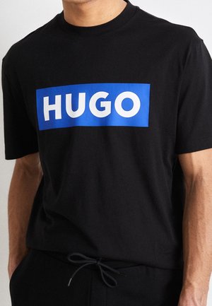 Mann trägt ein schwarzes T-Shirt mit einem blauen Rechteck und dem Wort "HUGO" in weißen Großbuchstaben auf der Brust.