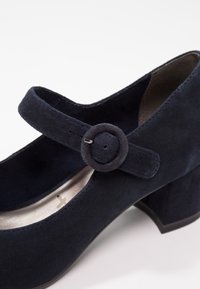 Chaussure Mary Jane en daim bleu marine avec bride à boucle arrondie et petit talon carré sur fond clair.