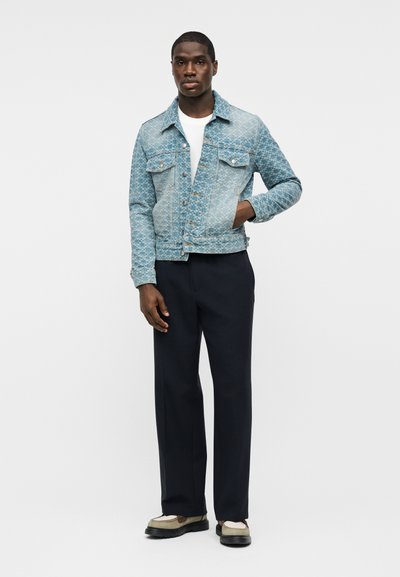 Άνδρας ντυμένος με πουκάμισο βαμβακερό τζιν σε ανοιχτό μπλε με σχέδιο, λευκό μπλουζάκι, μαύρο παντελόνι και μπεζ παπούτσια, στέκεται με το ένα χέρι στην τσέπη του μπουφάν.