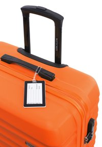 Oranžový tvrzený kufr s černou teleskopickou rukojetí a zipem. Obsahuje štítek na zavazadlo a texturovaný povrch s vertikálními drážkami.