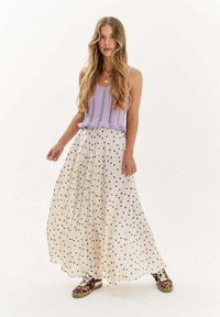Mladá žena s dlhými blond vlasmi, oblečená v levanduľovom pletenom topánke bez rukávov, bielej maxi sukni s bodkami a teniskách so vzorom leopardu, stojí.