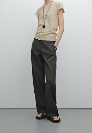 Personne portant un haut beige à manches courtes, un pantalon ample gris foncé, des sandales foncées, un long collier pendentif noir et un bracelet, debout avec une main dans la poche.