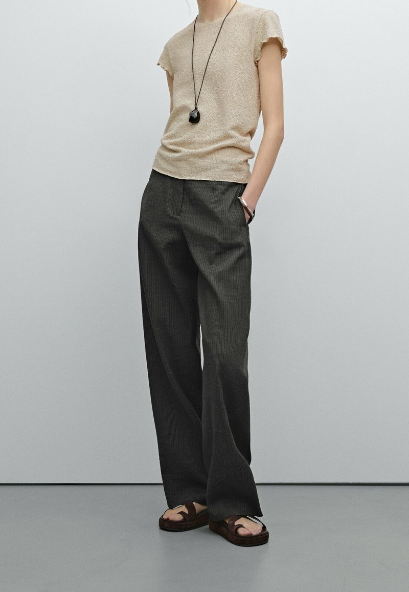 Personne portant un haut beige à manches courtes, un pantalon ample gris foncé, des sandales foncées, un long collier pendentif noir et un bracelet, debout avec une main dans la poche.
