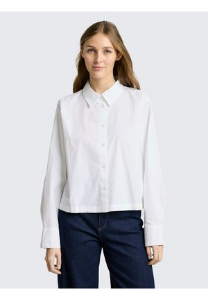 Junge Frau trägt ein locker sitzendes weißes Hemd mit breiten Manschetten und dunklen Jeans, steht vor einem schlichten hellgrauen Hintergrund.