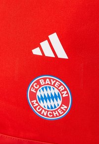 Rød stoff med hvit Adidas-logo og sirkulær FC Bayern München-emblemet med rød og blå diamantmønster. Glatt tekstur, sporty design.
