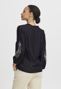 Blusa nera con maniche lunghe a sbuffo, caratterizzata da un intricato ricamo bianco sulle maniche. Realizzata in tessuto leggero e con scollatura rotonda.