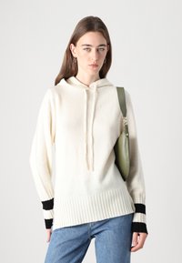 Krémová mikina s kapucňou a rebrovaným lemovaním, s čiernymi prúžkami na manžetách, doplnená zelenou taškou. Hladká textúra a neformálny dizajn.