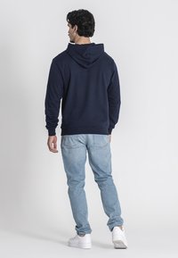 Sudadera azul marino con capucha ajustable, material de mezcla de algodón, combinada con jeans de mezclilla azul claro y zapatillas blancas. Costuras traseras visibles.