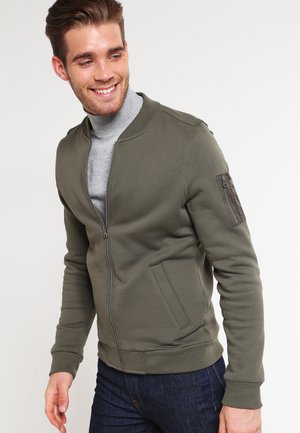 Olivengrøn zip-up jakke lavet af ribbet stof, med sidelommer og en lynlåslomme på venstre arm. Glat tekstur, tætsiddende design.