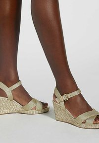 Sandales à talon compensé avec un design de bande tressée dorée, présentant une open toe, une boucle à la cheville et un talon compensé texturé.