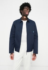 Chaqueta azul marino con cuello de botón, bolsillo frontal y un pequeño logo; camiseta blanca y pantalones de color claro. Textura suave.