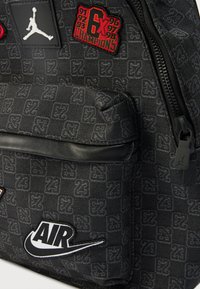 Black fabric backpack featuring the Jumpman logo, various patches including "6X Champions," and "AIR," with textured checkered background.