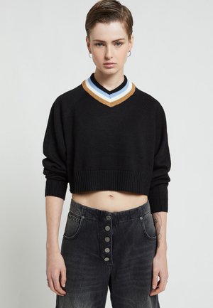 Giovane che indossa un maglione nero corto con scollo a V a righe e jeans scuri a vita alta con bottoni, in piedi davanti a uno sfondo semplice.