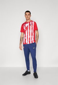 Piros és fehér csíkos sportpóló logóval, kék futónadrággal párosítva, amelyen sárga Nike pipa és oldalzseb található.