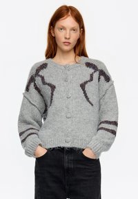 Szary sweter na guziki z kolorowymi akcentami i fakturowanym wzorem na rękawach, noszony na ciemnych dżinsach.