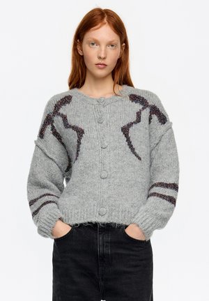 Szary sweter na guziki z kolorowymi akcentami i fakturowanym wzorem na rękawach, noszony na ciemnych dżinsach.