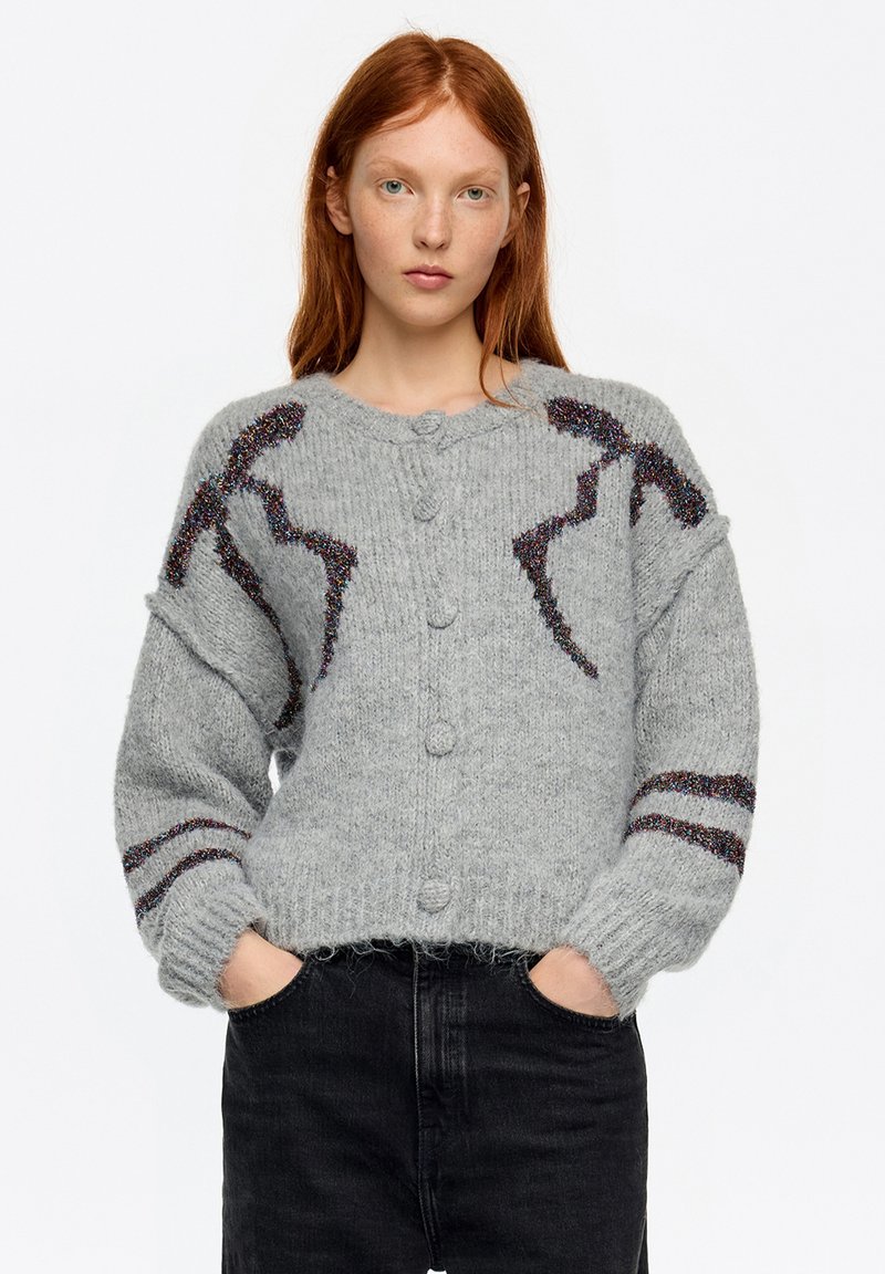 Szary sweter na guziki z kolorowymi akcentami i fakturowanym wzorem na rękawach, noszony na ciemnych dżinsach.