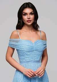 Ljusblå klänning med avskuren axel, samlad tyg och prickig textur. Tunna axelremmar och en figurnära bodice som leder till en flödande kjol. Silverarmband.