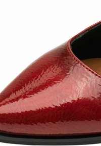 Chaussure en cuir verni rouge avec un bout pointu, présentant une texture lisse et un minimum de couture, reposant sur une semelle noire.