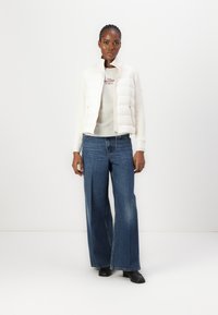Hvid puffjakke over en lysegrå sweater, parret med brede blå jeans og sorte sko. Glatte teksturer og enkelt design.