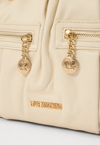 Μπεζ δερμάτινη τσάντα χειρός με δύο μπροστινές τσέπες με φερμουάρ, χρυσές λεπτομέρειες, αλυσιδωτή διακόσμηση και ανάγλυφο λογότυπο "LOVE MOSCHINO" στη βάση.