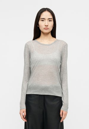 Νεαρή γυναίκα με μακριά σκούρα μαλλιά που φορά διαφανές ανοιχτό γκρι μπλουζάκι με μακριά μανίκια και μαύρο παντελόνι, στέκεται μπροστά σε απλό φόντο.