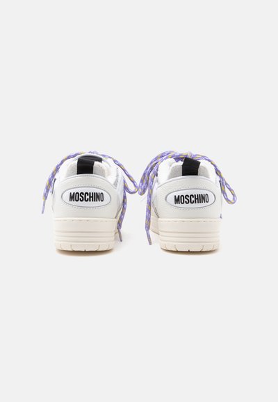 Λευκά αθλητικά παπούτσια Moschino με μωβ και κίτρινα κορδόνια, εμφανιζόμενα από πίσω σε απλό φόντο.