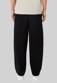Schwarze Jogginghose aus weichem Material mit lockerer Passform, ausgestattet mit zwei Gesäßtaschen und elastischem Saum. Weiße Sneaker runden den Look ab.