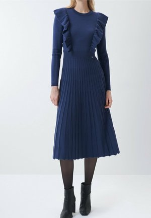 Navyblauwe gebreide jurk met lange mouwen, voorzien van ruches op de schouders en een geplisseerde rok. Het materiaal lijkt zacht met een textuurafwerking.