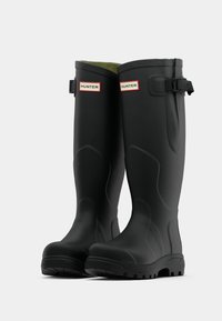 Botas de lluvia de goma negras con superficie texturizada, tobillo alto y correa ajustable. Presentan un interior verde y acentos de logo rojos.