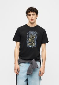 Νεαρός άνδρας με σγουρά μαλλιά, φορώντας μαύρο t-shirt Puma με πολύχρωμο αφηρημένο σχέδιο λιονταριού, ανοιχτό γαλάζια τζιν και δεμένη γκρι φούτερ γύρω από τη μέση.