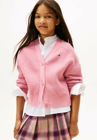 Cardigan en tricot rose avec un décolleté en V, doté de quatre boutons et de grandes poches. Porté sur une chemise blanche et assorti à une jupe à carreaux.
