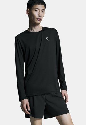 Junger Mann, der ein schwarzes langärmeliges Sportshirt und schwarze Shorts trägt, steht vor einem einfachen hellen Hintergrund.