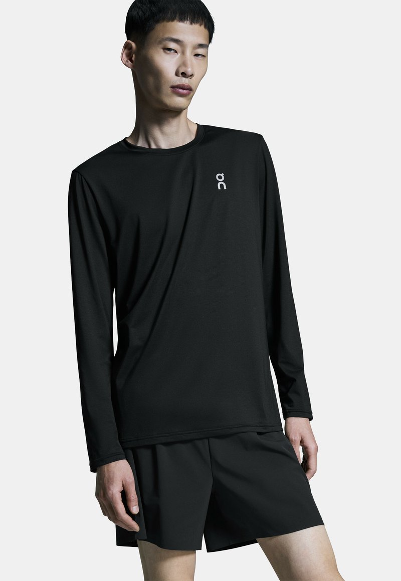 Jeune homme portant un t-shirt de sport noir à manches longues et un short noir, debout devant un fond clair uni.