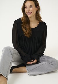 Blusa nera a maniche lunghe con dettagli arricciati e polsini a balze, abbinata a jeans grigi chiari. Il tessuto sembra leggero e leggermente trasparente.
