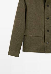 Olive green quilted jacket sleeve with black buttons and a rectangular flap pocket on a white background.