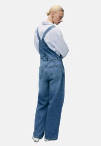 Denim kombinezone svijetloplave boje s podesivim trakama, širokim nogavicama, dva stražnja džepa i opuštenim krojem. Upareni s bijelom košuljom na gumbe.