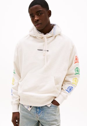 Cream-colored hoodie with drawstring, featuring printed logos in red, green, blue, and yellow on the right sleeve. Front kangaroo pocket.