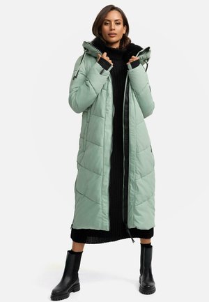 Lange, mintgroene pufferjas met een ritssluiting, capuchon en een zwart geribbelde trui eronder. Zwarte kniehoge laarzen maken de look compleet.