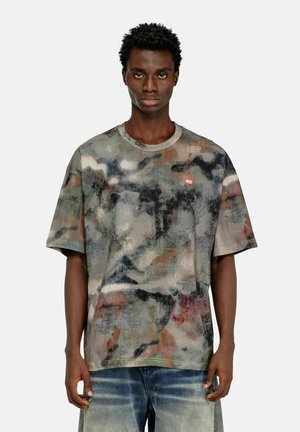 Giovane uomo con capelli corti e ricci che indossa una t-shirt larga multicolore stile mimetico e jeans blu sbiaditi, in piedi davanti a uno sfondo semplice.