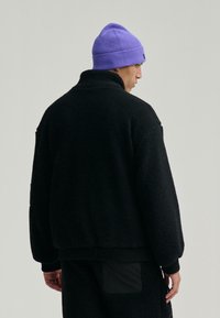 Czarna polarowa kurtka z wysokim kołnierzem i strukturalną powierzchnią; w połączeniu z jasnofioletowym, prążkowanym beanie; widok z tyłu ukazujący luźny krój.
