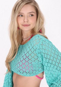Haut tricot turquoise avec motif en dentelle ouvert, manches larges et longueur cropped, associé à un bikini rose en dessous.