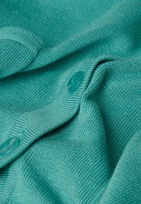 Türkisfarbener Strickstoff mitglatter Textur, versehen mit zwei passenden Knöpfen. Das Material fällt weich und betont seine Form.