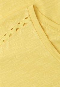 Tissu jaune avec un col en V et un détail de bordure festonnée le long de la couture de l'épaule.