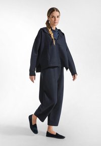 Marineblauwe hoodie met een voorzak, gedragen over een bijpassende marineblauwe trainingspak; zachte textuur, relaxte pasvorm en instap donkere schoenen.